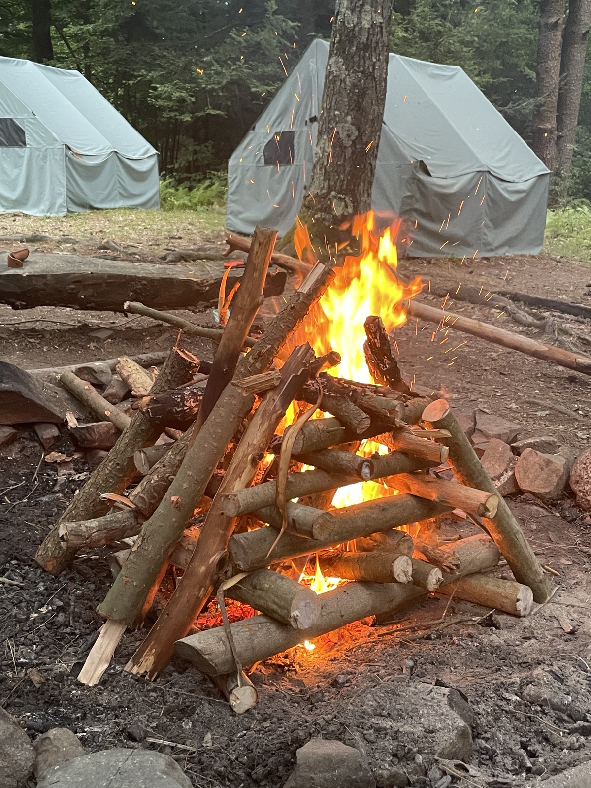 A teepee-style log fire crackles in front of platform tents at summer camp.